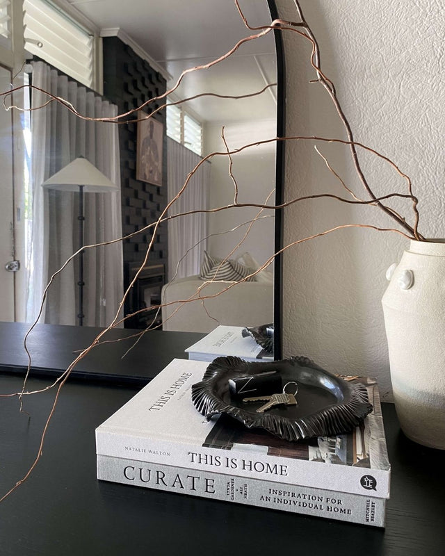 A handmade ceramic dish sits atop a stack of coffee table books on a console, with a minimalistic vase. A mirror reflects a styled lounge room with brick fireplace, sofa, artwork and contemporary lamp.