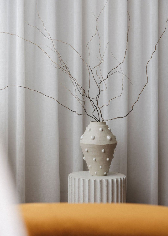 A large handmade ceramic vase with luxe pearl details sits on a plinth, styled with dried branches.