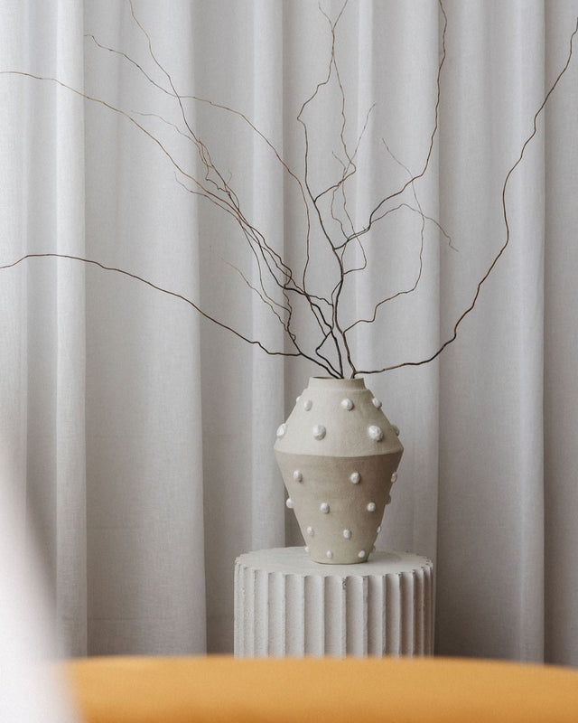 A large handmade ceramic vase with luxe pearl details sits on a plinth, styled with dried branches.